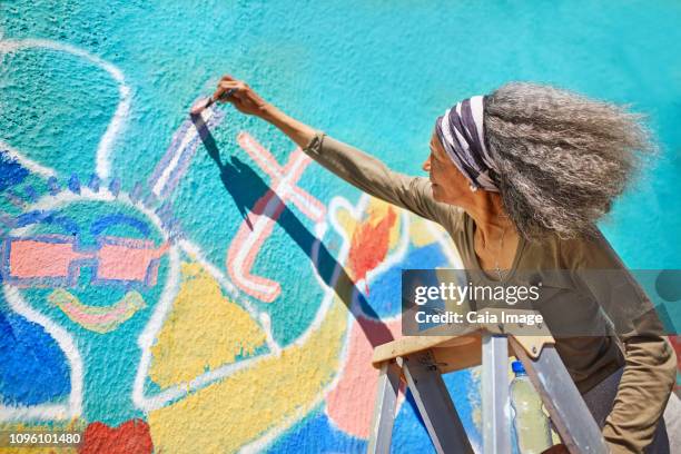 senior woman painting vibrant mural on sunny wall - wandmalerei stock-fotos und bilder