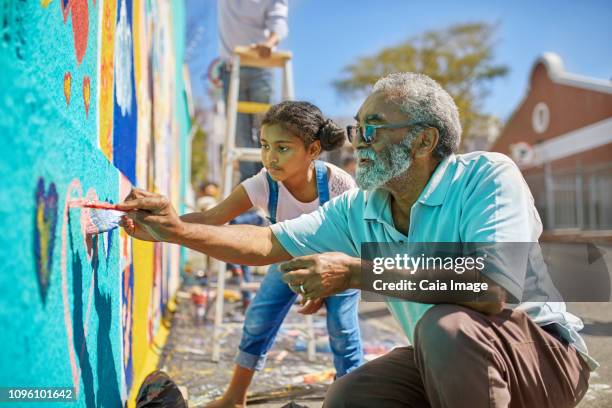 grandfather and granddaughter volunteers painting vibrant mural on sunny urban wall - murale foto e immagini stock