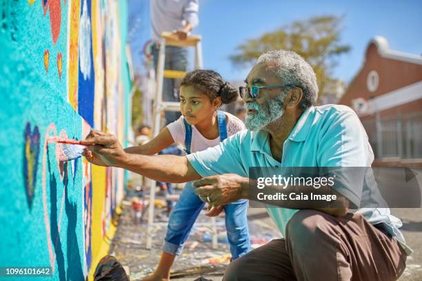 grandfather and granddaughter volunteers painting vibrant mural on sunny urban wall - muurschildering stockfoto's en -beelden