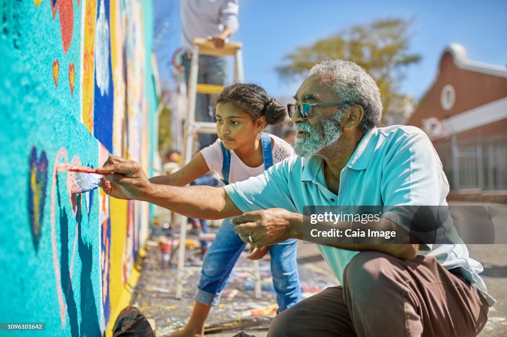 Grandfather and granddaughter volunteers painting vibrant mural on sunny urban wall