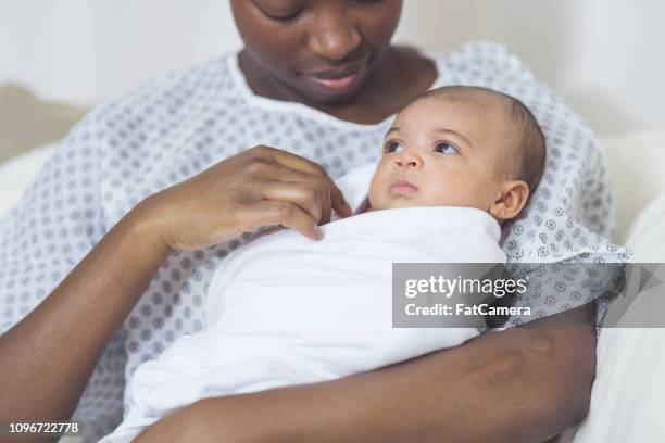 schöne afrikanische amerikanische mutter in ein krankenhemd hält ihr neugeborenes baby sanft an ihre brust - hospital gown stock-fotos und bilder