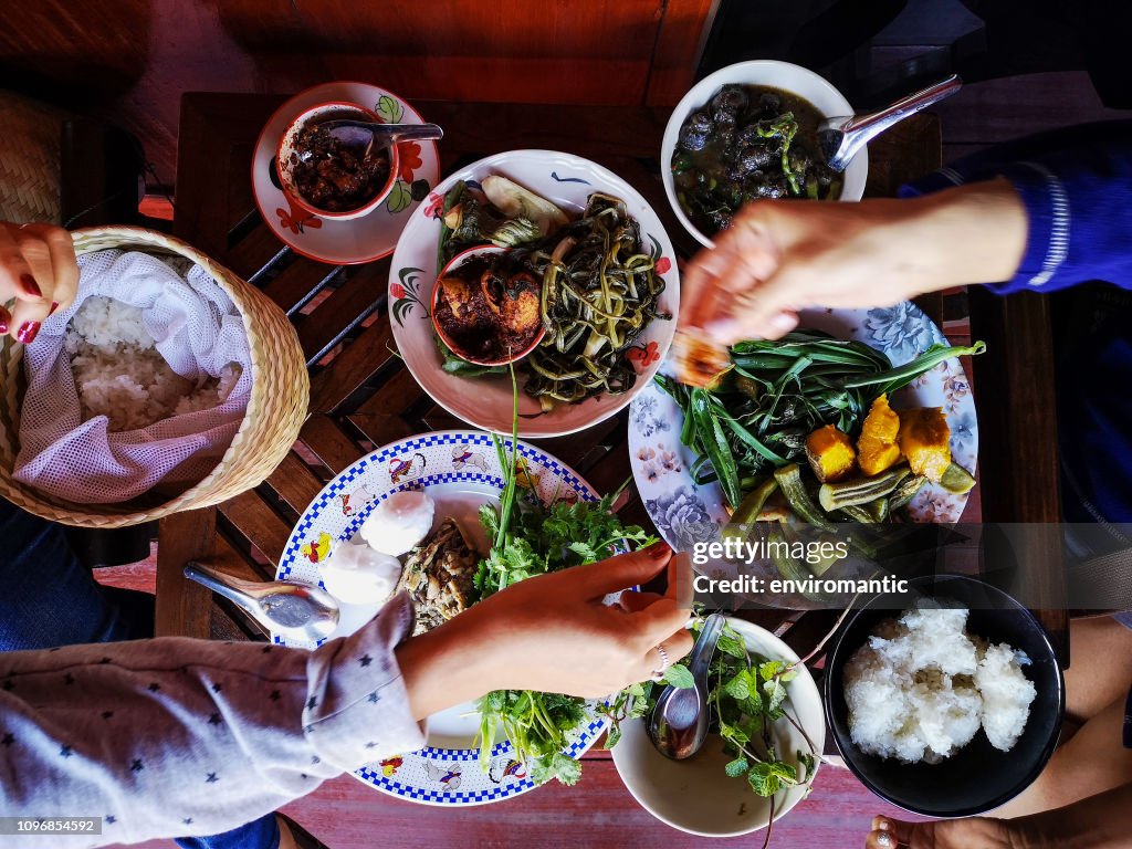 Two Thai women traditionally eating a selection of freshly cooked Northern Thai food of vegetables, soups and curries served in dishes on a wooden table, along with sticky rice, which is a food staple in Northern and Northeast Thailand.