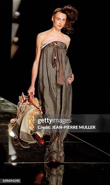 Model presents a high waisted grey linen pants with a crumpled tie as a belt, holding a basket containing a blanket, old newspaper, shoes... By...