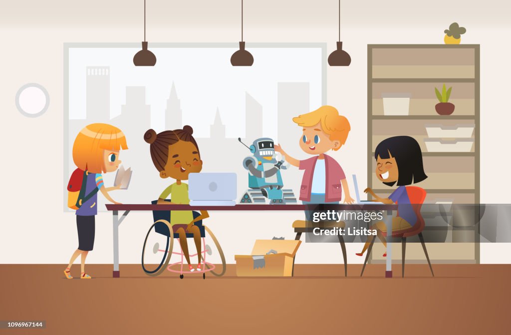 Disabled African American girl in wheelchair and other children standing around desk with laptops and robot and working on school project for programming lesson. Concept of inclusion at school.