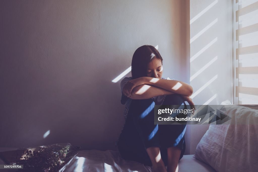亞洲婦女坐在床上抱著膝蓋。感到悲傷, 失望的愛情在黑暗的臥室和陽光從窗戶透過百葉窗。復古色調。