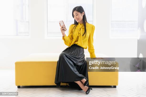 chinese woman on yellow bench with phone - bloco de cor imagens e fotografias de stock