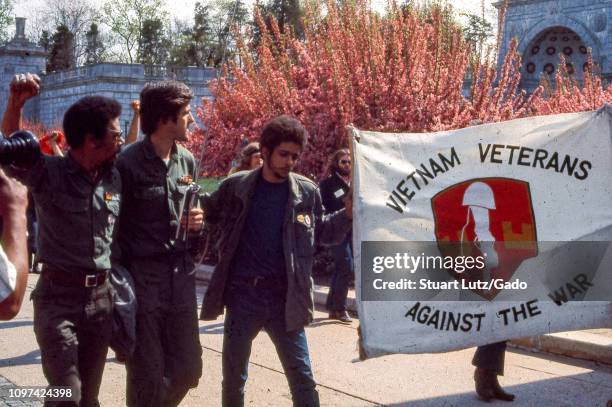 Landscape view of three men, including John Kerry, wearing military uniforms walking next to a banner with the text "Vietnam Veterans Against the...