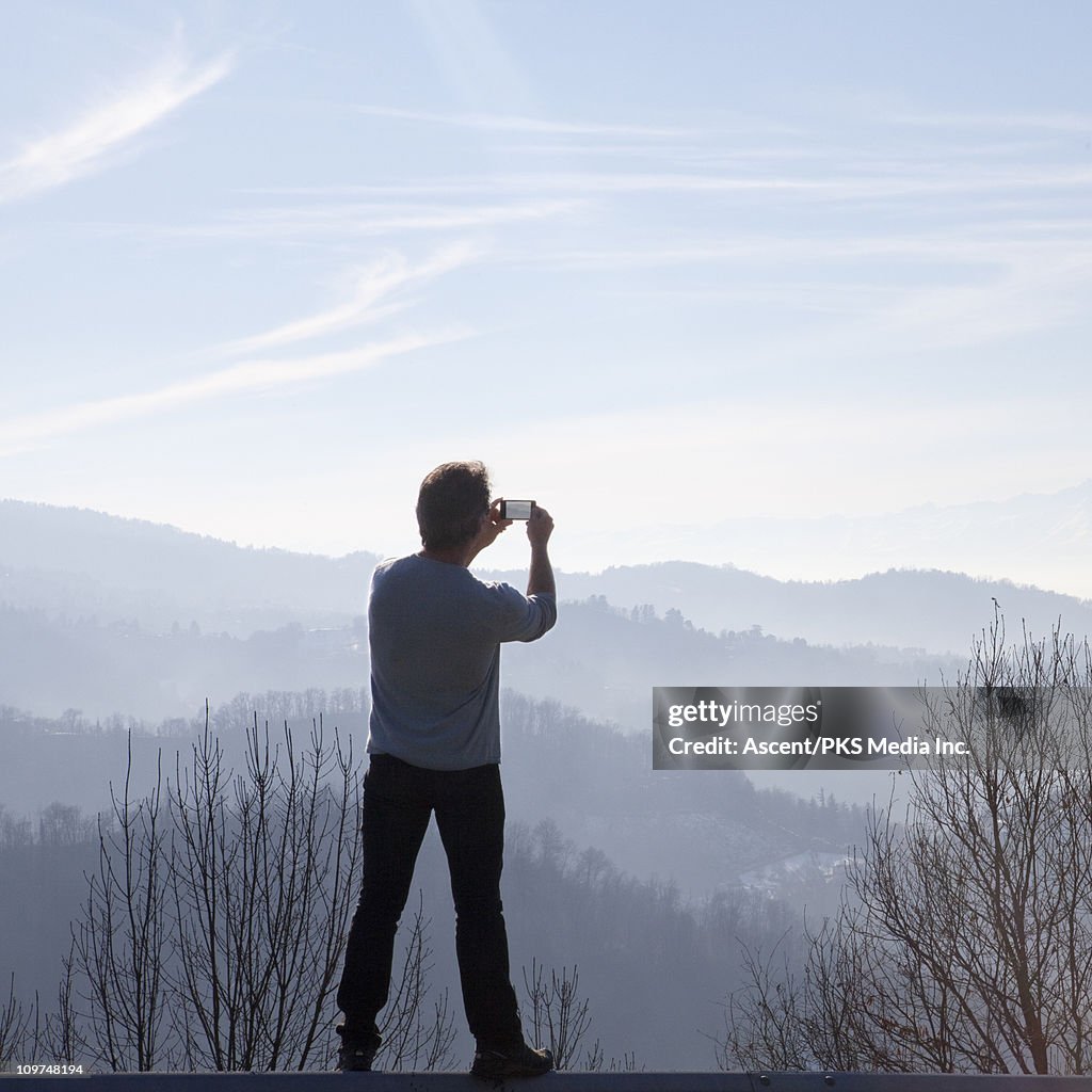 Man stands on fence rail, takes picture of hills