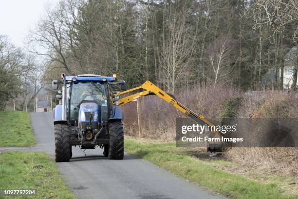 Tractor with hedge cutter on trimming the grass verge along a rural road, Cumbria, UK.