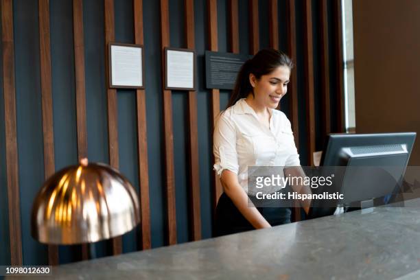 latin american hotel receptionist working behind counter smiling while looking at computer screen - hotel stock pictures, royalty-free photos & images