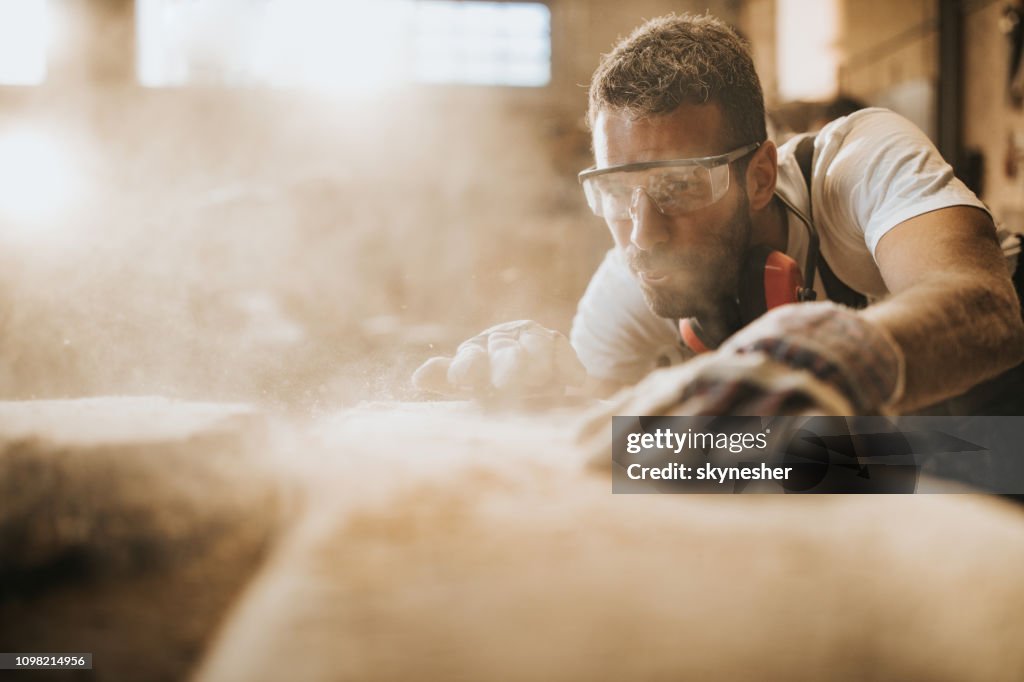 Carpenter blowing sawdust while working on a piece of wood in a workshop.