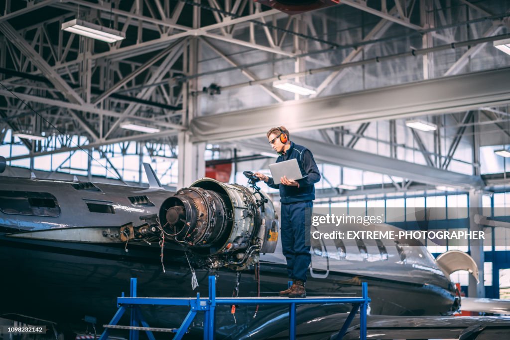Aircraft mechanic standing on a raised work platform, looking at a display of a probe inspection camera, trying to diagnose a fault in a jet engine of a small private airplane in a maintenance hangar