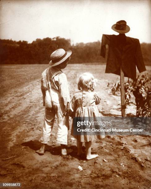 Two children, a boy and girl, stand and look up at a scarecrow, Cincinnati, Ohio, early 20th century.