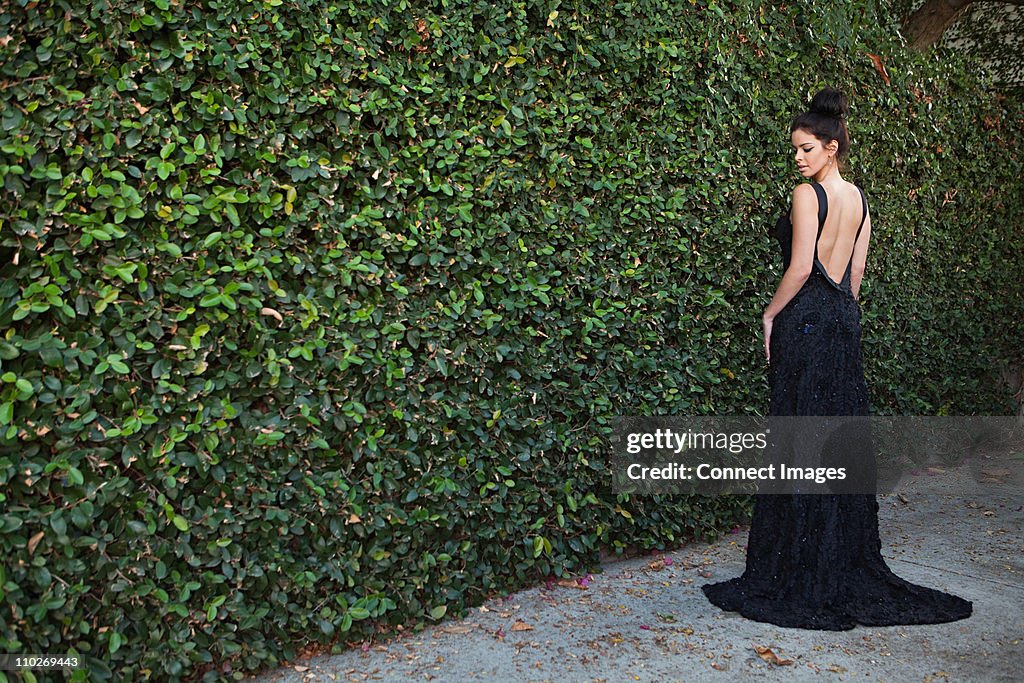 Young woman wearing black evening dress, standing by hedge