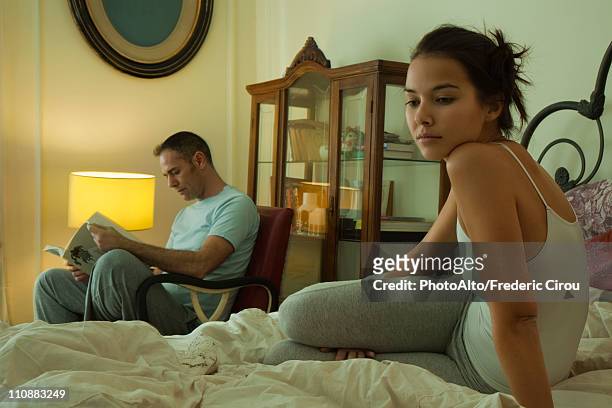 wife sitting on bed looking bored, husband reading in background - erotik stock-fotos und bilder