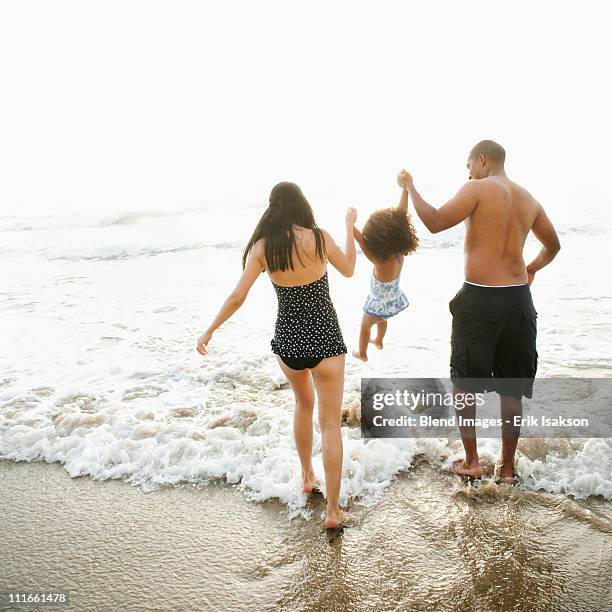 parents swinging daughter between them on beach - mother and child in water at beach stock pictures, royalty-free photos & images