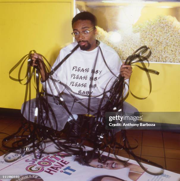 Portrait of African American film director Spike Lee, holding film reels, while crouching in front of a poster for his film 'She's Gotta Have It',...