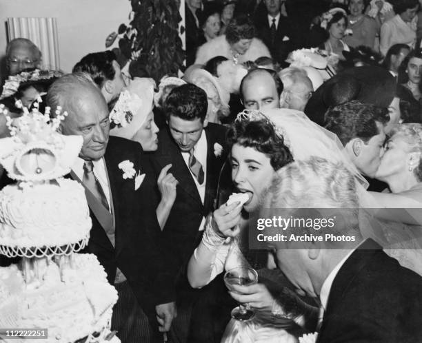 Elizabeth Taylor eating wedding cake at her wedding to Nicky Hilton, 6th May 1950. Her new father in law Conrad Hilton looks on and her father,...