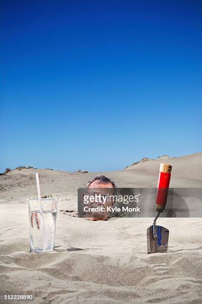 caucasian man buried in sand next to glass of water and trowel - futility stock pictures, royalty-free photos & images