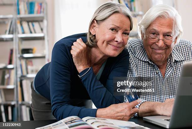 germany, wakendorf, senior woman with credit card and man using laptop - woman portrait kitchen laptop stock-fotos und bilder