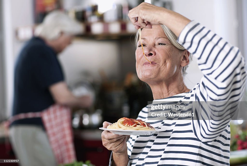 Germany, Wakendorf, Senior woman eating noodles, man cooking in background