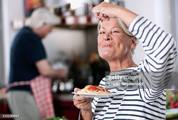 germany, wakendorf, senior woman eating noodles, man cooking in background - senior adult eating stock pictures, royalty-free photos & images