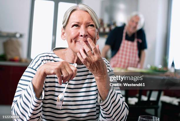 germany, wakendorf, senior woman tasting noodles, man cooking in background - flavoring stock-fotos und bilder