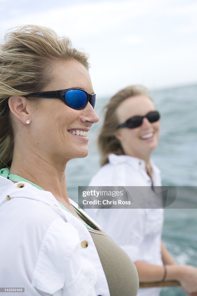 Two women with sunglasses smile with the ocean in the background.