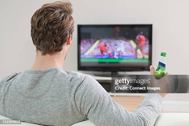 young man watching football on television with beer bottle - man cave stock pictures, royalty-free photos & images