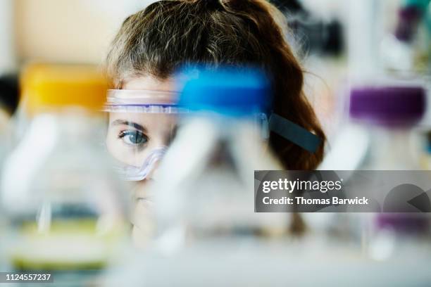 view through bottles of solution of female scientist wearing safety goggles in laboratory - kleine scherptediepte stockfoto's en -beelden