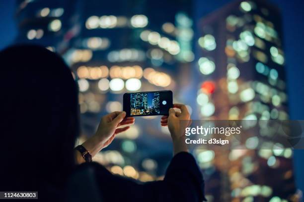 woman capturing the prosperity of city night scene with smartphone against illuminated cityscape - woman capturing city night stockfoto's en -beelden