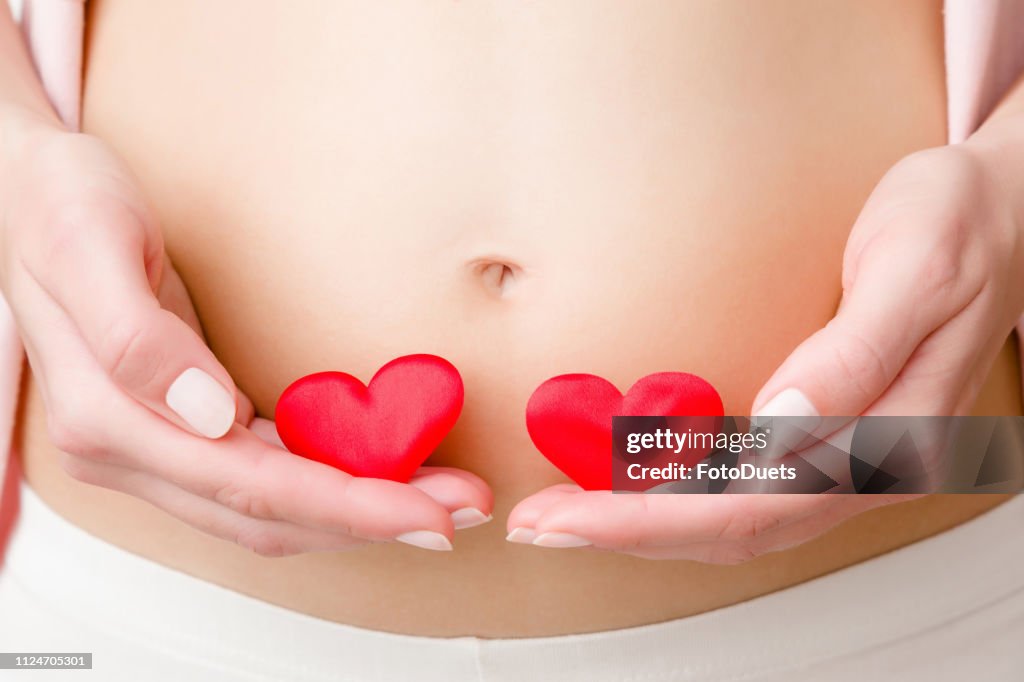 Young woman's hands holding two red bright hearts on naked belly. Emotional loving moment in pregnancy time. Twins babies expectation. Love, happiness and safety concept. Close up. Front view.