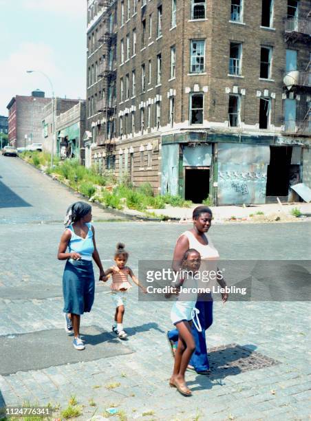 Wide view of an unidentified group of women and children as they cross a street in front of a bombed-out tenement building in New York City's...