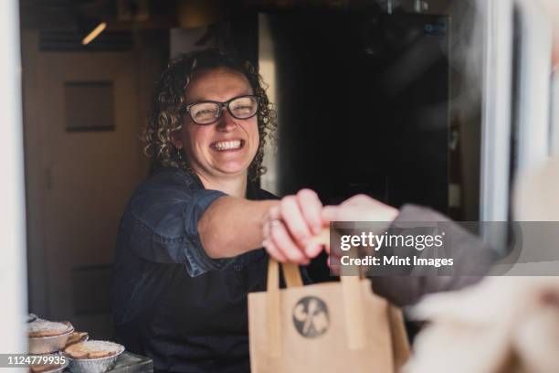 smiling woman wearing glasses handing  brown paper shopping bag through window of bakery. - homegrown produce stock pictures, royalty-free photos & images