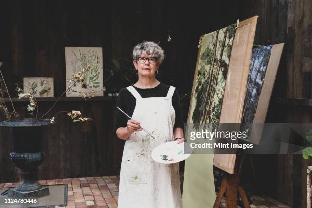 senior woman wearing glasses, black top and apron standing in studio, working on painting of trees in forest. - artist portrait stock pictures, royalty-free photos & images