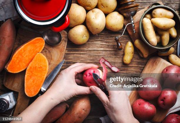 womans hand peeling a potato, top view. variety of raw uncooked organic potatoes: red, white, sweet and fingers potatoes over wooden background. - zoete aardappel stockfoto's en -beelden