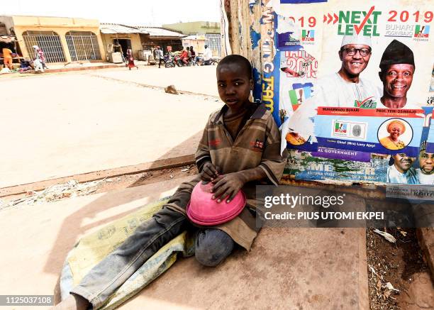 Young beggar sit with a bowl beside poster of candidate of the ruling All Progressives Congress President Mohammadu Buhari and Vice President Yemi...