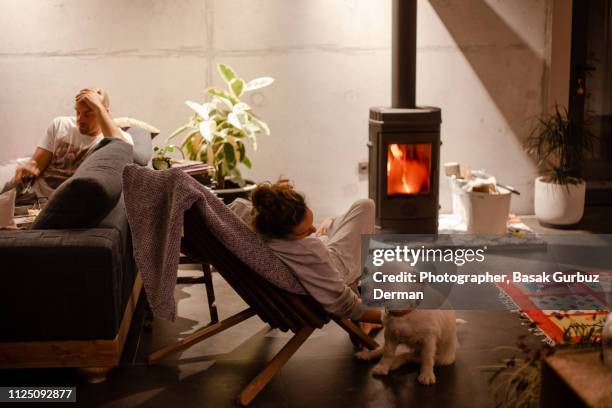 a woman petting her dog in front of a stove in a cozy living room while a man sitting in a sofa - winter dog stock pictures, royalty-free photos & images