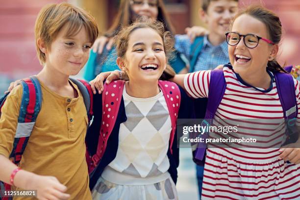 escolares están caminando al frente de la escuela y ríen juntos - first day of school fotografías e imágenes de stock