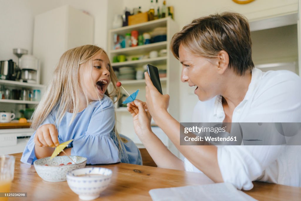 Happy mother and daughter having fun at table at home taking smartphone picture