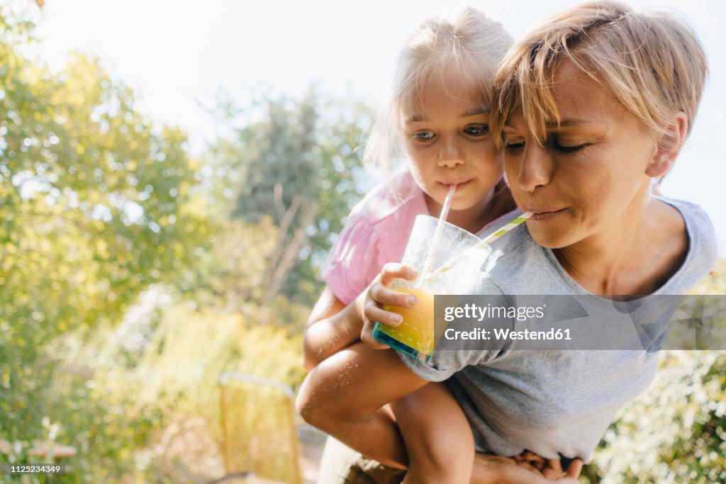 Mother carrying daughter piggyback in garden drinking a smoothie