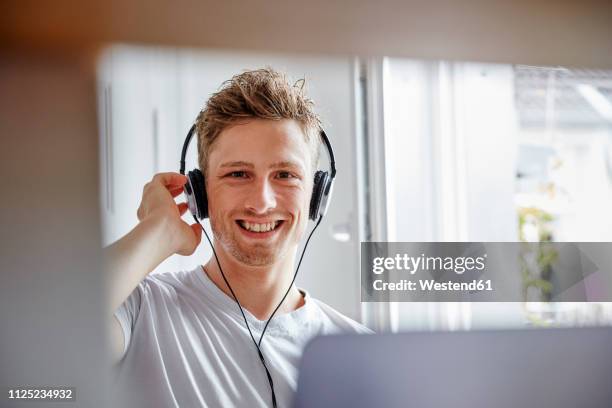 portrait of smiling young man at home listening to music with headphones - bearded blonde man stock-fotos und bilder