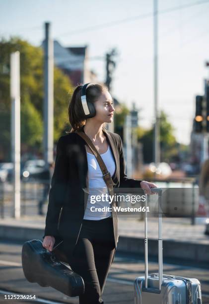 young woman with headphones, suitcase and violin case at tram station - borsa a tracolla foto e immagini stock