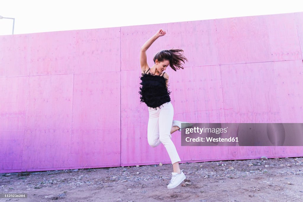 Exuberant young woman jumping in front of pink wall