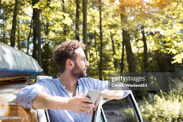 young man with cell phone at car in forest - auto mann stock-fotos und bilder