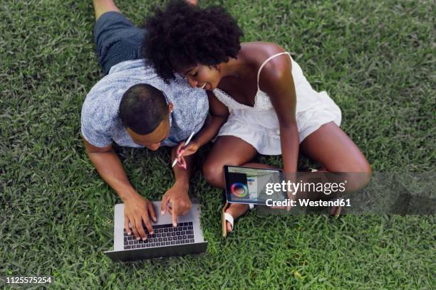 young couple using tablet and laptop on lawn in a park - tablet paar sommer stock-fotos und bilder