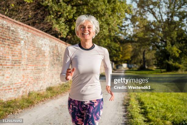 happy senior woman running along brick wall in a park - corredora de footing fotografías e imágenes de stock
