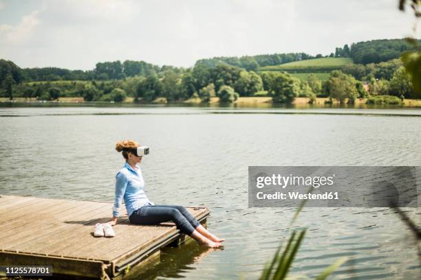 woman with vr glasses sitting on jetty at a lake with feet in water - water glasses ストックフォトと画像
