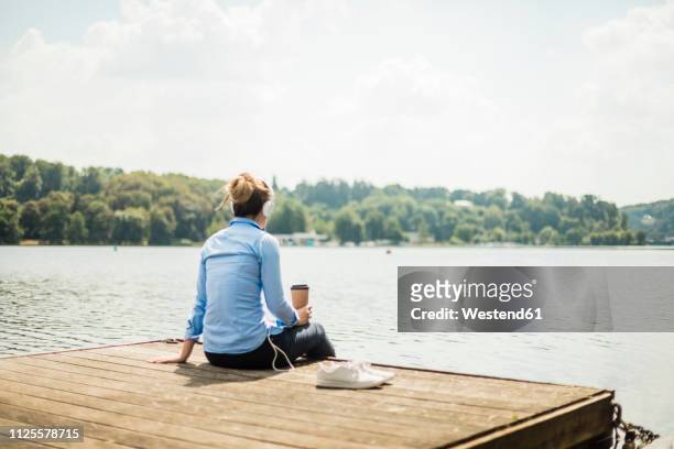 woman sitting on jetty at a lake with headphones and takeaway coffee - essen ruhrgebiet stock-fotos und bilder