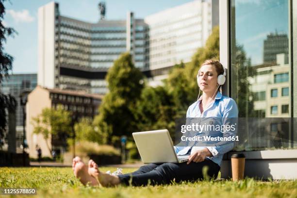 woman wearing headphones sitting on urban meadow using laptop - arbeiten outdoor stadt laptop stock-fotos und bilder